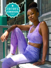 Photo of Lukafit founder sitting in a baseball dugout wearing Lukafit standout sports bra and squat-proof leggings. Her bra and leggings are both lavender ombre color with our signature print of women doing different active yoga and workout poses. She is looking directly at the camera with a faint smile. One of her arms is resting on her raised knee.